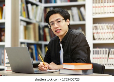 Working On Research Project. Portrait Of Asian Male Student Using Laptop At Library, Writing In Notepad, Looking At Camera