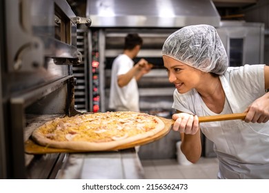 Working In Pizza Restaurant. Female Chef In White Uniform And Hairnet Putting Pizza In The Oven For Baking Process.