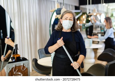 Working Process In Beauty Salon During Pandemic Situation. Elderly Female Hairstylist In Protective Mask Cleaning Work Space, Sweeping Floor