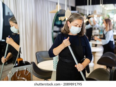 Working Process In Beauty Salon During Pandemic Situation. Elderly Female Hairstylist In Protective Mask Cleaning Work Space, Sweeping Floor