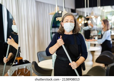 Working Process In Beauty Salon During Pandemic Situation. Elderly Female Hairstylist In Protective Mask Cleaning Work Space, Sweeping Floor