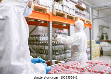 Working Process In Food Factory Warehouse: Group Of Workers Wearing Coveralls And Safety Masks Wrapped Up In Loading Goods, One Of Them Holding Digital Tablet In Hands