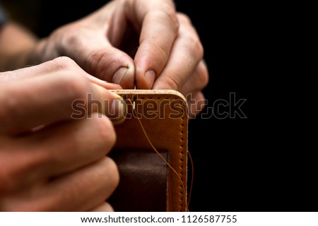 Working process of the leather belt in the leather workshop. Man holding crafting tool and working. He is sewing to make a walet. Tanner in old tannery. Wooden table background
