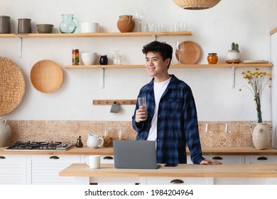 Working remotely, freelancing and taking care of health during self-isolation. Smiling young asian guy holding glass of water in modern kitchen interior with laptop and cup on table, free space