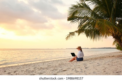 Working remotely on seashore. Young successful woman female freelancer in straw hat working on laptop while sitting on tropical beach at sunset, full length. Distance work concept - Powered by Shutterstock