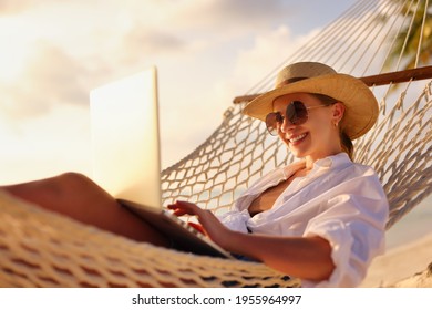 Working Remotely. Side View Of Young Happy Woman, Female Freelancer In Straw Hat And Sunglasses Working On Laptop While Relaxing In The Hammock On The Beach At Sunset. Distance Job During Vacation