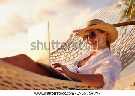 Working remotely. Side view of young happy woman, female freelancer in straw hat and sunglasses working on laptop while relaxing in the hammock on the beach at sunset. Distance job during vacation