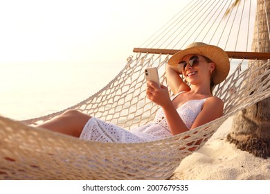 Working remotely. Young happy woman, female freelancer in straw hat and sunglasses working on smartphone while relaxing in the hammock on the beach at sunset. Distance job during vacation - Powered by Shutterstock