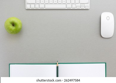 Working Space In Grey-green. Creative Office Style Composition With Green Apple, Computer Keyboard And Mouse, Big Opened Note Pad With Green Pencil, Located On Grey Wooden Desk. Top View.