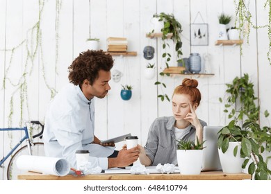 Working in team. Caring Afro American architect sitting on desk with rolls of blueprints, tools and crumpled papers, offering coffee to his unhappy woman coworker, trying to support and reassure her - Powered by Shutterstock