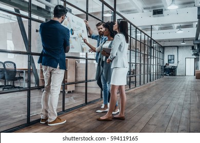 Working As A Team. Full Length Of Young Modern People In Smart Casual Wear Planning Business Strategy While Young Woman Pointing At Large Paper In The Office Hallway