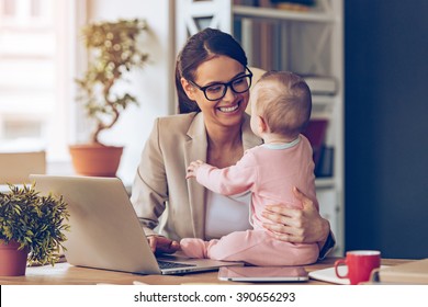 Working together is so fun! Cheerful young beautiful businesswoman looking at her baby girl with smile while sitting at her working place - Shutterstock ID 390656293
