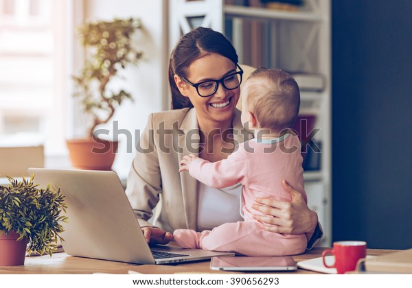 Working together is so fun! Cheerful young\
beautiful businesswoman looking at her baby girl with smile while\
sitting at her working\
place