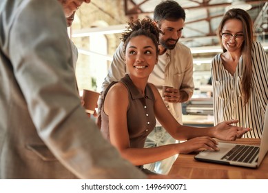 Working Together. Young And Cheerful Afro American Woman Using Laptop And Discussing Something With Colleagues While Sitting In The Modern Office. Teamwork. Office Life