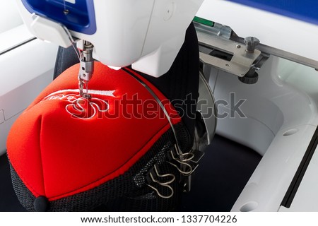 Working white embroidery machine embroidering logo on red and black sport cap, close up picture
