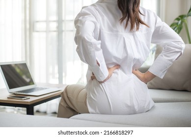 Working woman sitting on the sofa in the living room at home She used both hands to press down on the lower back. She is suffering from back pain from sitting for a long time. - Shutterstock ID 2166818297