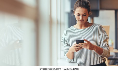 Working Woman Standing Beside A Window Holding Mobile Phone. Woman Entrepreneur Taking A Break From Work Checking Her Cell Phone.