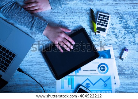 A working womans hand at night at a table with laptop, tablet and phone.
