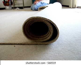 A Workman In Blue Jeans And T-shirt Kneels Next To A Roll Of Carpet Ready To Be Cut And Installed