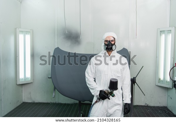 Workman in protective suit and\
respirator holding aerograph and looking at camera in\
garage