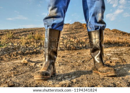 A workman stands with his black, dirty rubber boots on a gravel road in front of a pile of earth in the light of the evening sun.
