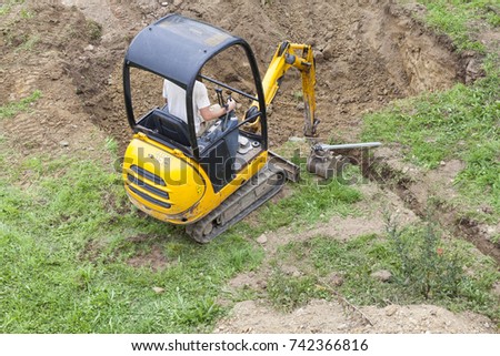 Workman using a mini digger to excavate  a hole for a swimming pool in a garden lawn with green grass viewed from above.
