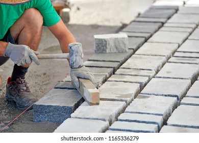 A workman's gloved hands use a hammer to place stone pavers. Worker creating pavement using cobblestone blocks and granite stones. - Shutterstock ID 1165336279