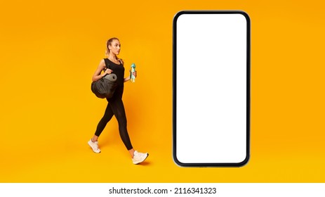 Workout App. Fitness Lady Walking To Huge Cellphone Screen Carrying Sporty Bag And Bottle Of Water Over Yellow Studio Background. Gym Mobile Offer Or Application Concept. Panorama, Mockup