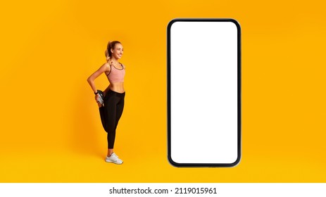 Workout Application. Fitness Female Exercising Near Big Phone With Blank Screen Stretching Leg Over Yellow Background In Studio. Great Mobile Offer, Online Gym App Concept. Mockup, Panorama