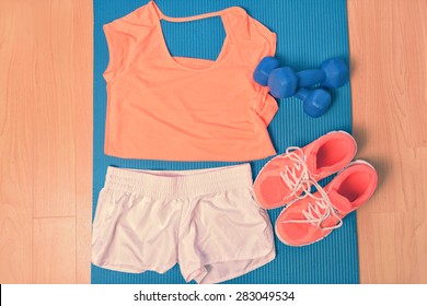 Workout Clothes - Fitness Outfit And Running Shoes. Overhead Of Clothing Ready For Lifting Weights At The Gym Or At Home, Laying On A Yoga Mat On The Floor. Matching Orange T-shirt And Sneakers.