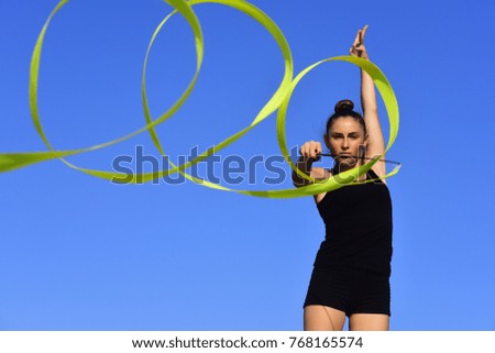 Workout of girl on blue sky background. Gymnastic school and energy. Sport and success. Flexibility in acrobatics and fitness health. Woman gymnast in black sportswear with green ribbon.
