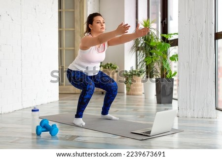Workout at home - middle aged woman performs squat exercise while looking into a laptop monitor. She uses an online tutorial.