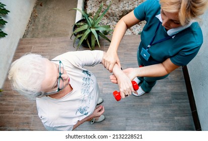 Workout Old Arm Muscles With Help Of Physiotherapist. Senior Female Exercise With Red Dumbbells On Physiotherapy Session. Helping To Rehab And Recovery Of Senior Woman Hands