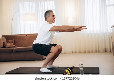 Workout Time Strong Senior Man Doing Squat Exercises At Home In His Light Living Room. Healthy Lifestyle, Wellbeing And Activity Concept. Black And White Sportswear. Active And Happy Old Age.