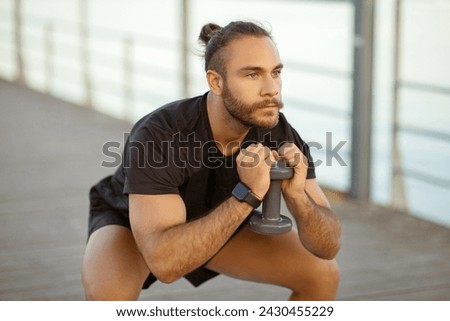 Workout with weights. Determined athletic bodybuilder guy squatting gripping dumbbell tightly, exercising outdoor at sea pier, looking aside. Concept of strength and sport discipline