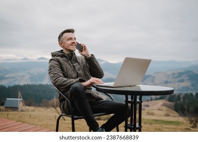 Workplace in country on backyard house in morning. Man talking on phone outdoors. Male work on a laptop sitting table with great view mountains. Concept remote work or freelance lifestyle. Internet 5G