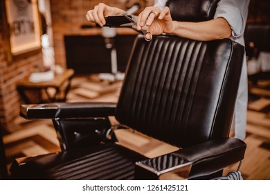 Workplace Of Hairdresser. Stylish Vintage Barber Chair Interior. Master Holds Scissors And Comb In Hands Leaning On His Back