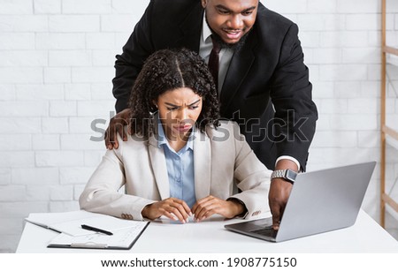 Workplace harassment concept. Boss touching his young attractive female assistant while she working on laptop at company office. Young woman suffering from offensive sexual behaviour