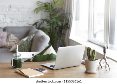 Workplace Of Modern Freelancer Leading Healthy Lifestyle: Open Generic Laptop Computer, Cactus In Pot, Smart Phone, Copybook And Glass Of Green Smoothie On Wooden Desk In Stylish Home Office Interior