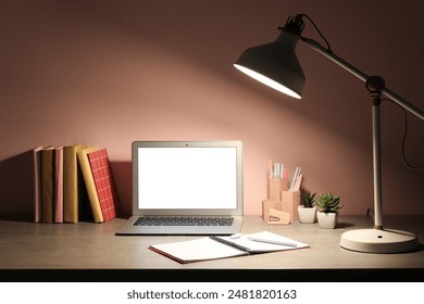 Workplace with modern laptop, notebook and glowing lamp near pink wall