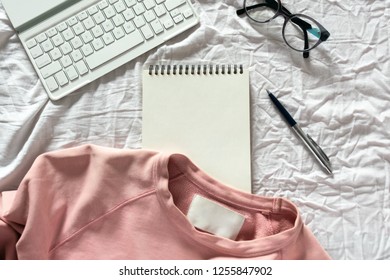 Workplace On Bed. Keyboard, Notebook, Glasses And Pink Sweatshirt On A White Crumpled Sheet. Flatlay, Top View