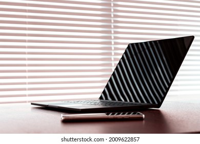 Workplace with opened laptop computer and cellphone liying on table above window with blinds. Natural sunset light with deep shadows. Toned close up image with soft selective focus