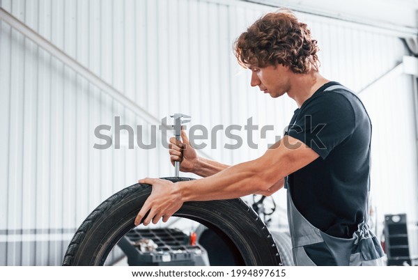 Works with tire. Adult man in grey colored uniform\
is in the automobile\
salon.