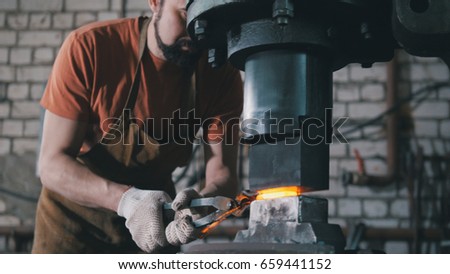Workshop of a blacksmith. A man puts a forged piece under an automatic hammer