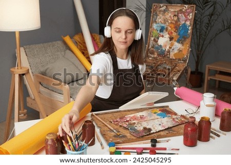 Workshop for creative minds. Showcasing artistic talent. Watercolor techniques and craft. Brown haired woman artist drawing with paints sitting in studio workshop and listening to music in headphones