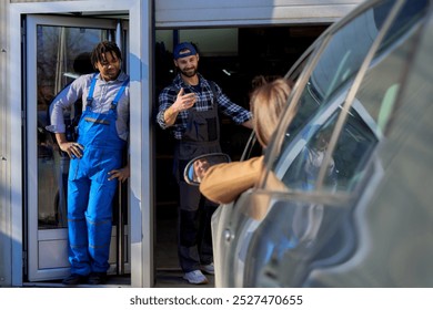 At the workshop entrance, a diverse team mechanic guides a woman on how to enter the garage. She peers through the window, seeking the right path.  - Powered by Shutterstock