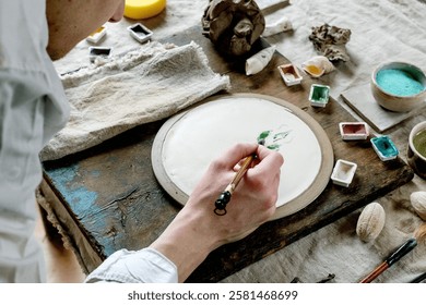 Workspace in ceramic studio, paints and brushes for underglaze painting. Young man's hands painting white plate. Branches, natural decor, linen tablecloth, daylight. Workshop idea.