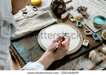 Workspace in ceramic studio, paints and brushes for underglaze painting. Young man's hands painting white plate. Branches, natural decor, linen tablecloth, daylight. Workshop idea.