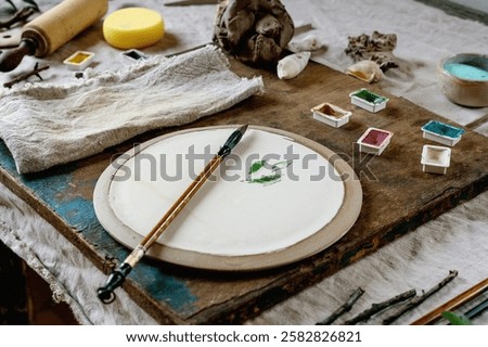 Workspace in ceramic studio, paints and brushes for underglaze painting. White plate, branches, natural decor elements, linen tablecloth. Daylight, idea for workshop.