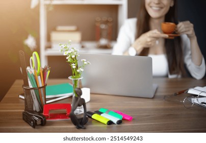 Workspace featuring colorful office supplies, a laptop, and vase of flowers, with a smiling woman holding a cup of coffee in the background. The bright, organized setting exudes creativity - Powered by Shutterstock